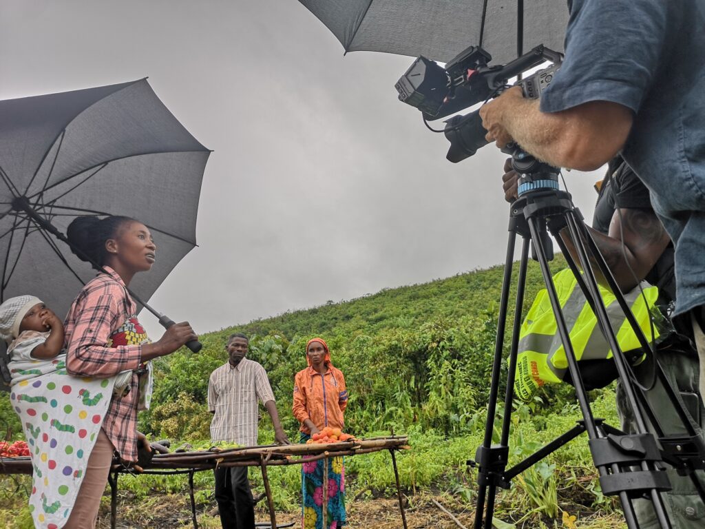 Kvinne med barn på ryggen som holder en paraply, to småbønder i bakgrunnen og et filmkamera