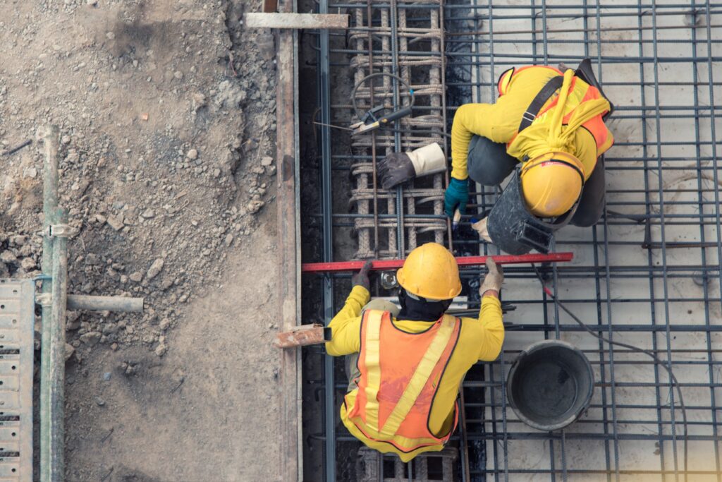 Two workers in orange overalls and yellow hard hats work on laying rebar at a construction site.