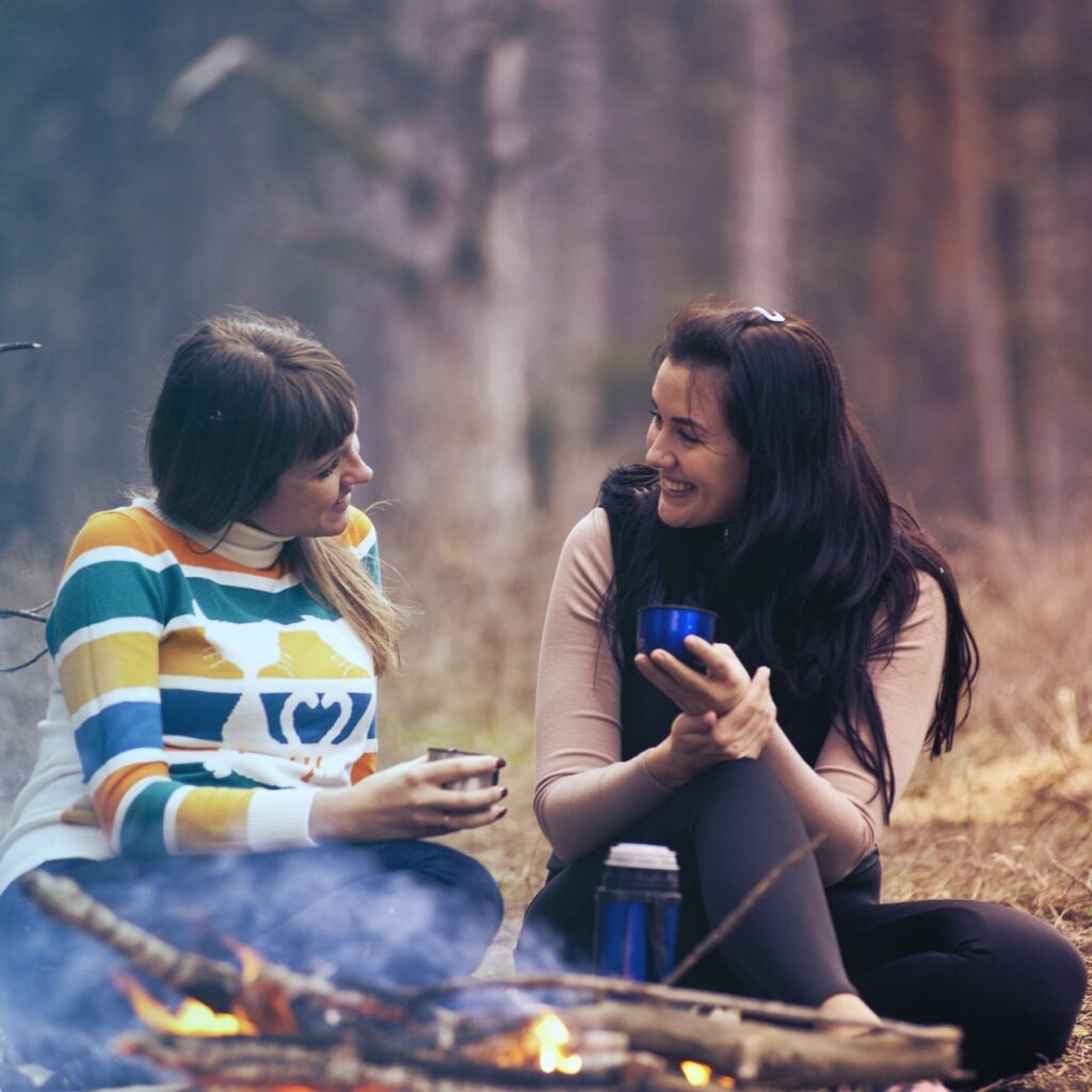 Dos mujeres de paseo por el bosque sentadas frente al fuego, cada una con una taza termo