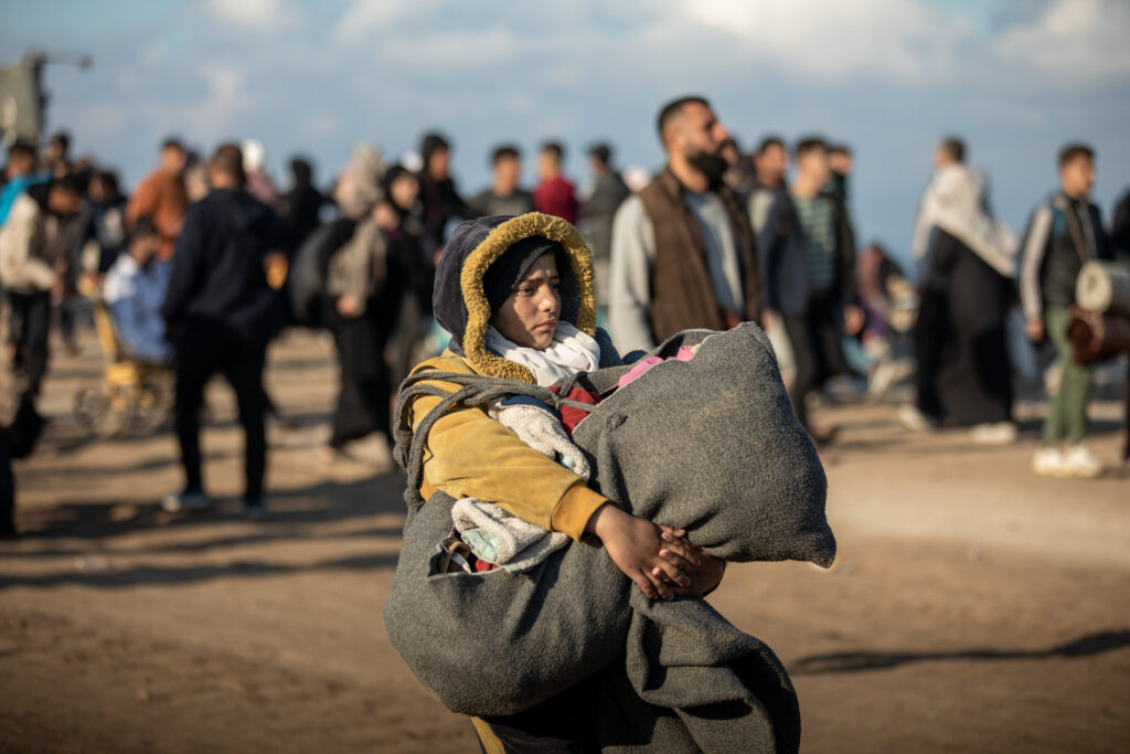 Ung gutt i Gaza, han bærer på alle sine eiendelen i en bag, og har på seg en tjukk jakke med hette. Det er mange mennesker i bakgrunnen som går samme vei som han. 