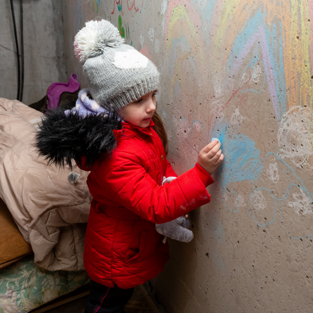 Ung jente fra Ukraina som tegner med kritt på en betongvegg i en bunker. Hun har på seg en rød jakke, grå lue og holder en kosebamse. 