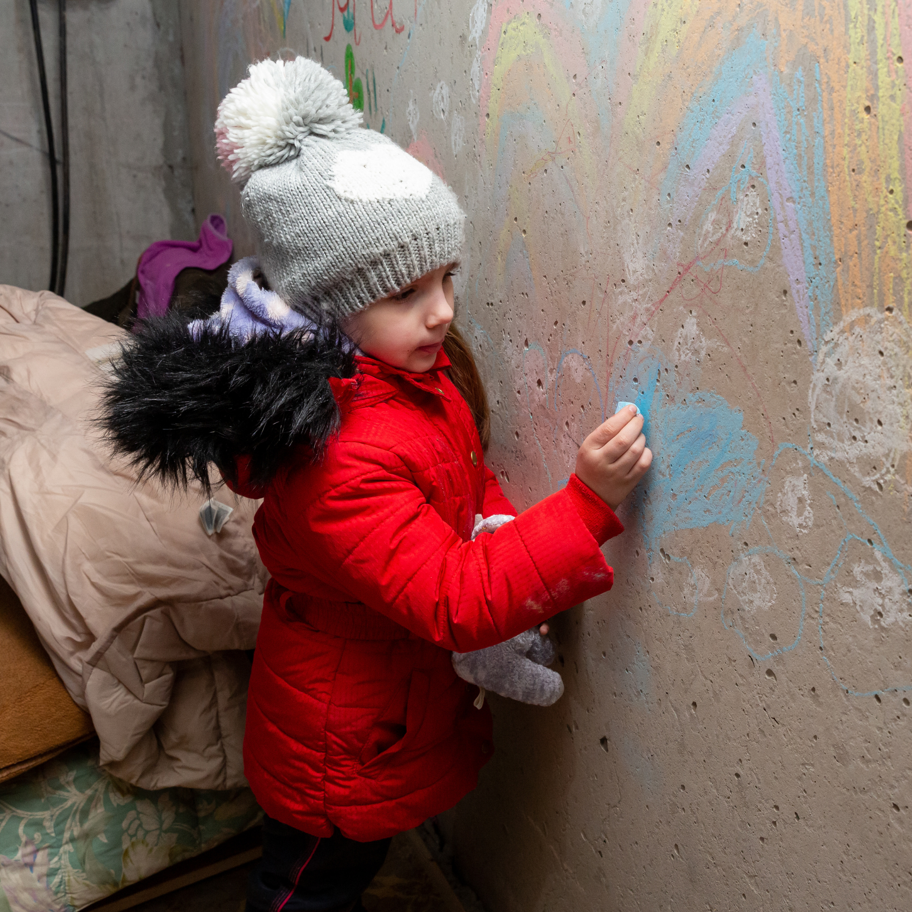Ung jente fra Ukraina som tegner med kritt på en betongvegg i en kjeller. Hun har på seg en rød jakke, grå lue og holder en kosebamse. (Foto: Vladimir Gerasimov, CRS)