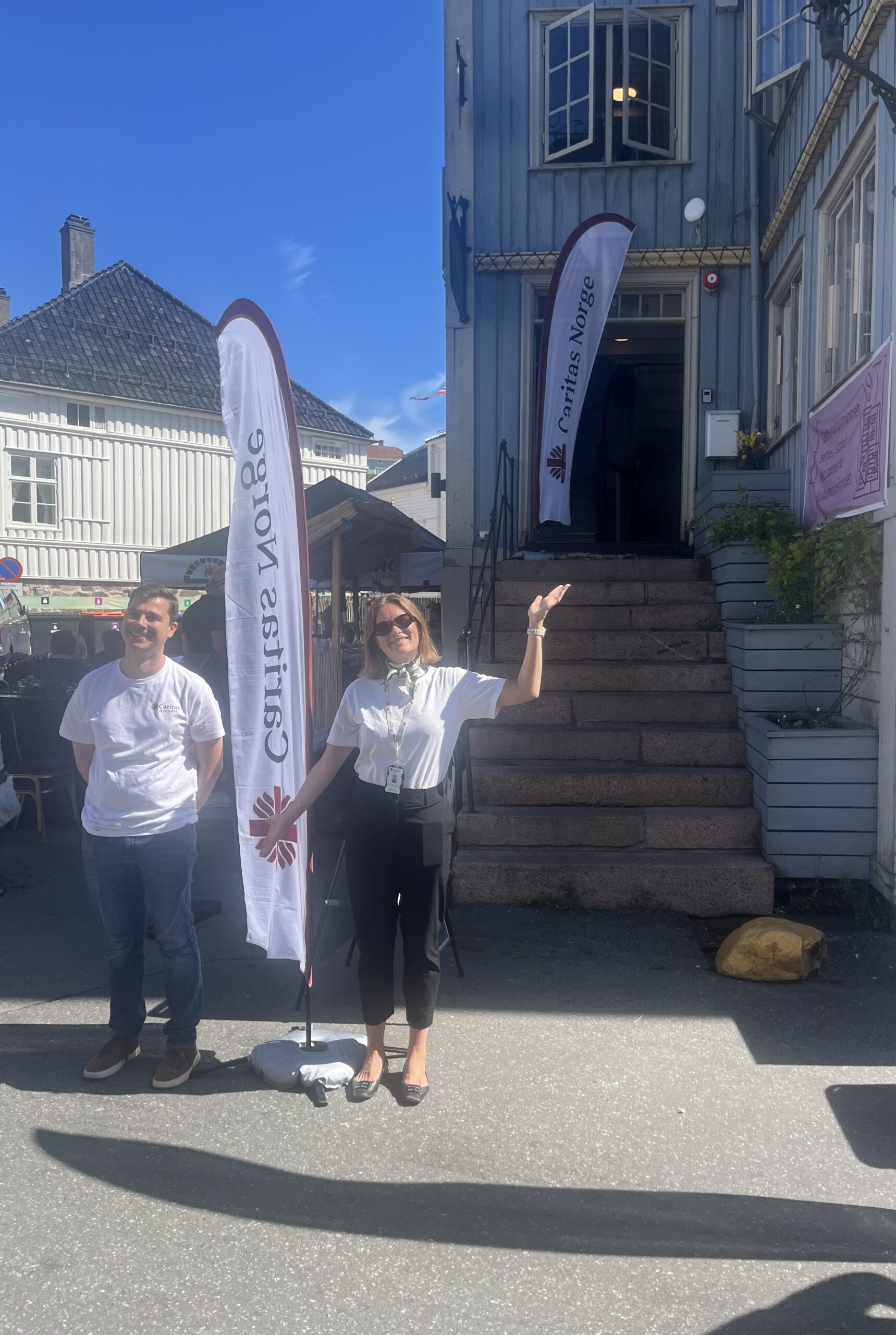 To ansatte i Caritas og en Caritas-vimpel utenfor Arendal Kunstforening sine lokaler hvor vi under Arendalsuka 2026 vil invitere til debatt.